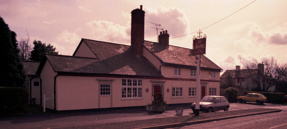 A two-storey building with a one-storey extension to the left side and a tall chimney in between, with a pub sign and a white Renault 5 car in front