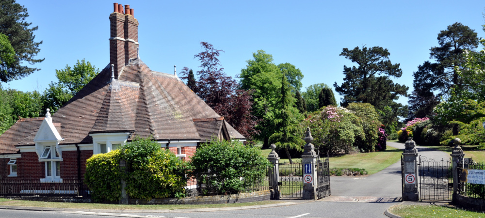 A small brick building with an elaborate roof and two tall chinmeys, next to an elaborate open gate, with trees and shrubs in the background