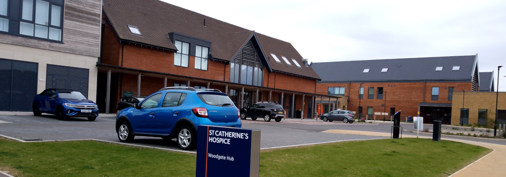 Modern two-storey buildings on two sides of a car park, with a sign reading: St Catherine's Hospice, Woodgate Hub