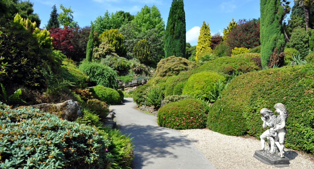 An elaborate series of hedges, shrubs and trees, with a path in the foreground and in the lower left-hand corner a small statue of two figures