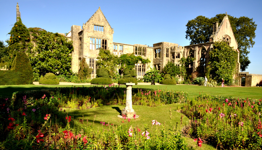 Flowers, most of them red, surrounding a sundial on a neat lawn, with the empty facade of a country house in the background