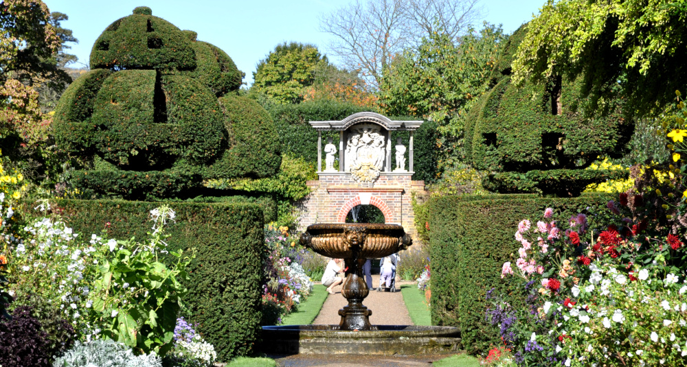 An ornate fountain in front of a sculpted wall, surrounded by hedges and flowers