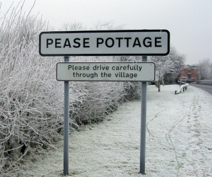 Road sign in a wintry landscape, reading: Pease Pottage - please drive carefully through the village