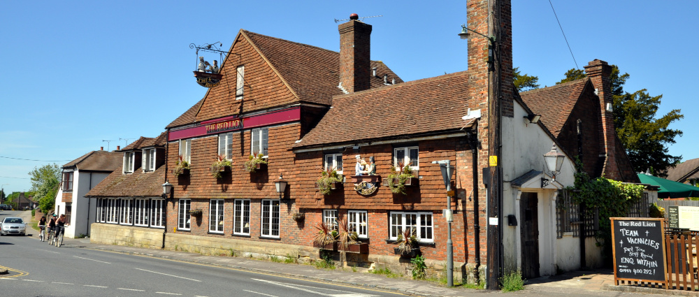 A wide, old, two-storey building close to a road, with a red sign reading: The Red Lion