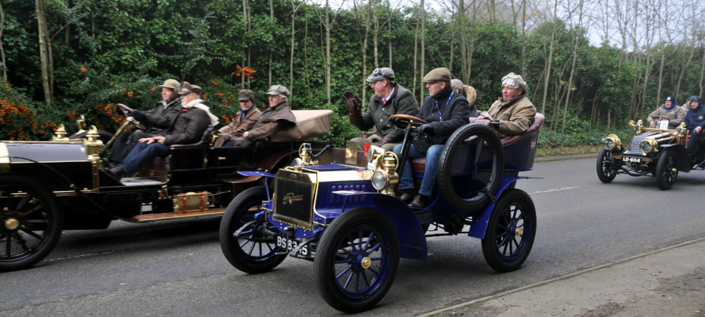 Three veteran cars close together on a narrow road with trees in the background
