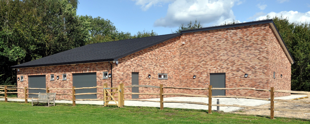 A wide, single-storey brick building with large doors and small windows; a fence in front and trees to the sides