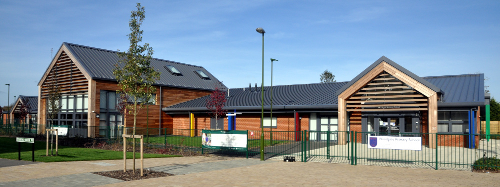 To the left, a modern two-storey building with wood cladding; to the right, a modern one-storey brick building; in front of both, a green metal fence containiing a sign reading: Woodgate Primary School