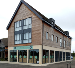 A modern two-storey building with a steep roof, large windows on the ground floor, and wood cladding above, with a green sign reading: Woodgate Community Shop