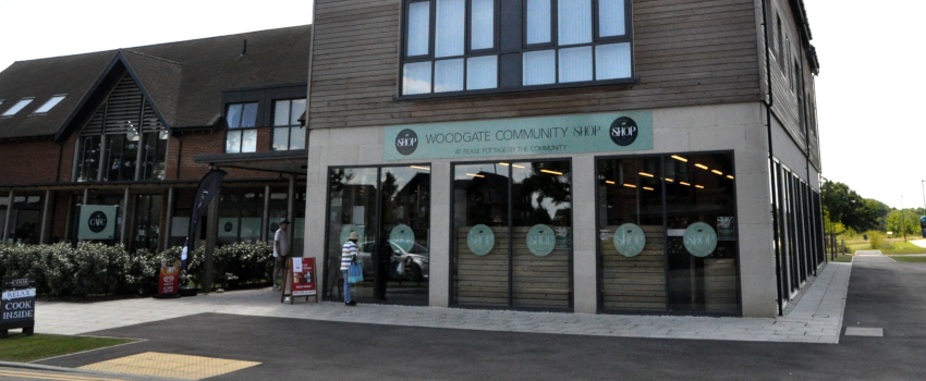 Two people in front of a modern building with large glass windows, and a green sign reading: Woodgate Community Shop
